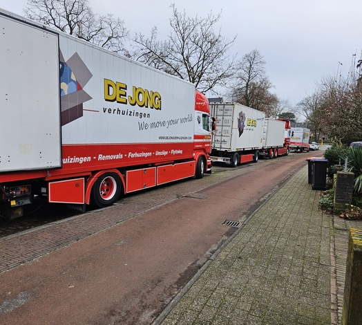 Meerdere verhuiswagens van De Jong Verhuizingen staan opgesteld in een woonwijk tijdens een verhuizing.